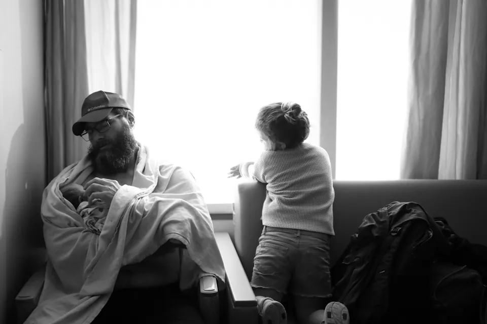 A father holds his newborn by the window while big sister looks out, black and white birth photography by Jenn Miller